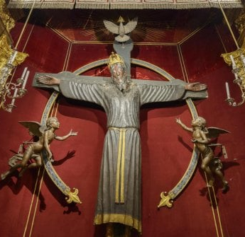 Photo of a crucifix ringed by two angels and with a dove above; the figure on the cross is robed, bearded, and wears a crown.