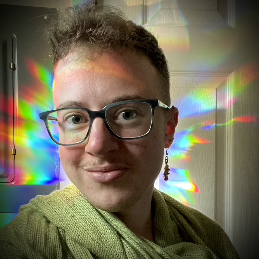 Photo of Avery Arden, a white person with short brown hair, light mustache, glasses, and dangly earring smiling at the viewer. Rainbow light shines behind their head