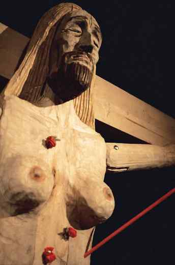 Detail of a the torso and head of wooden statue, as well as one arm stretched in crucifixion. The figure has long hair and a beard; the torso has breasts and an arrow embedded under the left breast.